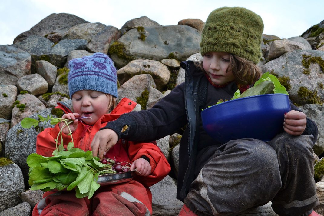 Barn sitter vid en stenmur i vinteroveraller och äter grönsaker. 