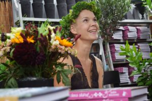 Sara sitter vid ett bord med blommor och tittar uppåt med ett stort leende, iförd eb krans av grönsaker.