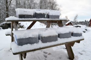 Plastbackar fyllda med jord och snö står uppradade på ett utebord i ett snöigt landskap med röda stugor i bakgrunden.