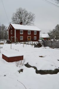 Skillnadens trädgård i vinterlandskap, längst fram syns flera odlingslådor och i bakgrunden det röda trähuset.