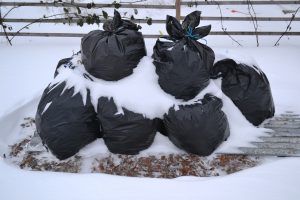 Sex fyllda, svarta sopsäckar står halvt täckta av snö ute i en vintrig köksträdgård.