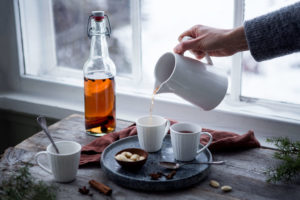 Framför ett ljust fönster syns någon hälla upp glögg ur en vit kanna i vite muggar. Kryddor och nötter pryder träbordet och brickan de står på.