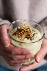 Närbild av två händer som håller i ett glas med en cremevit puré med gula, röda och bruna strimmor ovanpå.