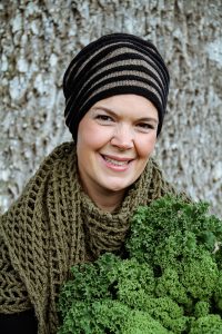 Porträttbild av Sara med randig stickad mössa, mörkgrön stickad halsduk och ett fång med grönkål i famnen.