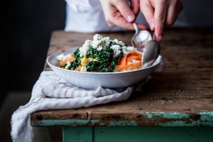På kanten av ett bord står en tallrik på en handduk. På tallriken ligger en färgglad sallad och två händer tar upp maten med skedar.