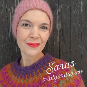 Selfie av Sara i rosa mössa och stickad ulltröja i många färger framför en gråbrun trävägg.