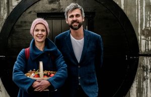 Sara och Johannes står bredvid varandra framför en svartmålad dörr, Sar ha en fruktkorg i famnen.