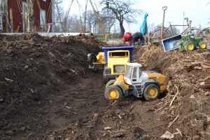 Leksakstraktorer och lastbilar står i en djupt, grävd ränna i jorden i köksträdgården.