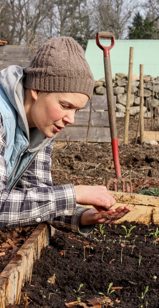 Sara lutar sig över en odlingslåda med frö i handen. Hon har en rutig skjorta, mössa och ser ut att berätta någonting.