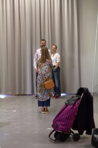 Sara och Johannes står framför ett stort, grått draperi. Johannes fru Therese tar kort på dem med ryggen till. I förgrunden syns en lila Dramaten-väska med svarta prickar på, där det hänger en kavaj.