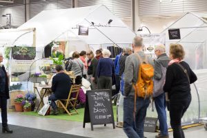 Gjordnäras monter på Nordiska Trädgårdar. Två stora tunnelväxthus och ett vägg-växthus ramar in en liten plats med bord och stolar.
