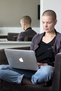 Sara sitter i en grå fåtölj på hotellrummet,med en grå dator i knät och ser allvarlig ut.