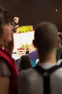 Vi ser Sara suddig bakifrån bredvid en scen på Nordiska Trädgårdar där Lena Israelsson föreläser.