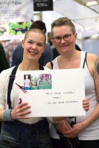 Sara i vit tröja och svarta hängslen står bredvid Veronica i vitt linne. Framför sig håller de en stor ihopbunden bok med ett foto av Sara och texten "Tack för vinsten! Från I ur och skur Hudiksvall"