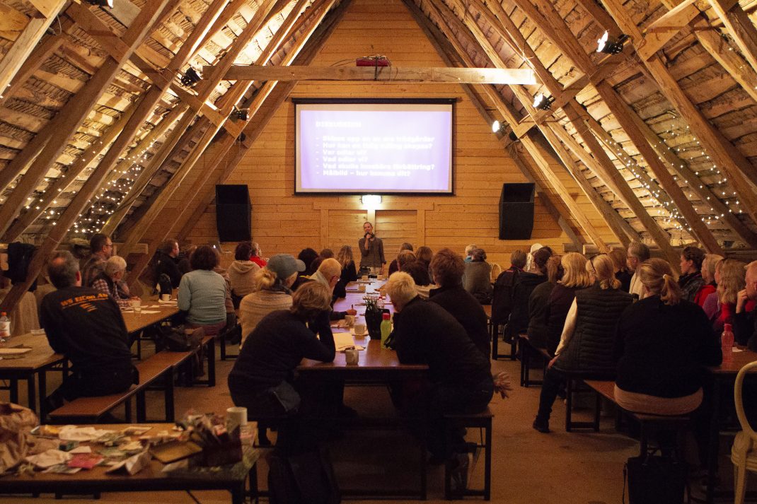 Översikt över loftet på Rinakby Gård, ett stort rum i trä med synliga balkar och ett skarpt sluttande tak. I rummet står långbord fulla med folk.