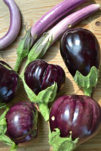 Två olika sorter av aubergine ligger på ett bord; avlånga och runda. De är mörkt lila och glänser i skalet.