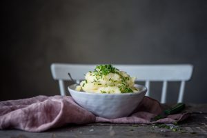 En skål med ljusgul puré av palsternacka står på ett träbord. Purén är toppad med gräslök och under ligger en syrenlila linneduk.