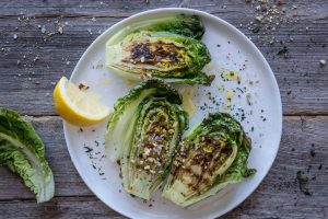 På ett vitt, runt fat ligger tre sallatshalvor, vackert grillade, med en citron bredvid.