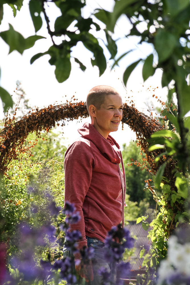 Sara står i röd tröja i solen mellan buskar och blommor.