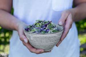 En person i vita kläder håller en skål i stenkeramik, fylld med coleslaw i grönt och rött.