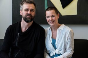 Johannes och Sara sitter i en soffa och tittar in i kameran, Sara ler.