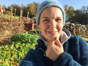 Selfie på Sara Bäckmo som tittar in glatt i kameran framför en höstig köksträdgård.