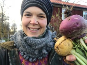 Sara håller upp några rovor framför sitt röda hus.
