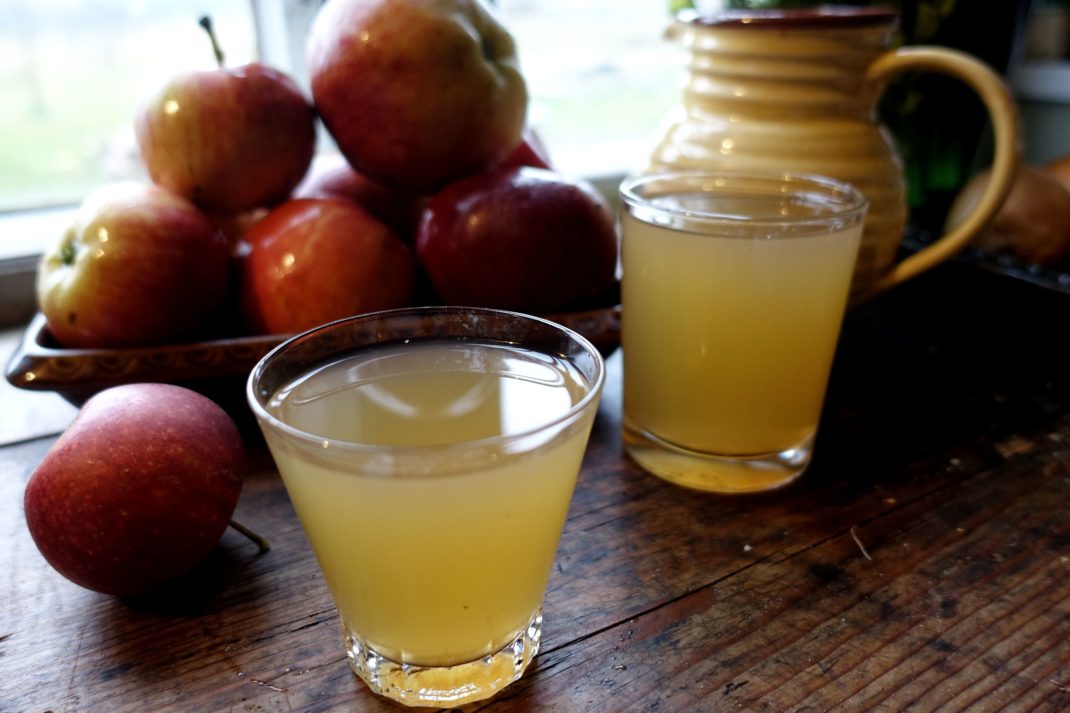 Två glas med äppelmust på ett träbord med en skål äpplen i bakgrunden. 