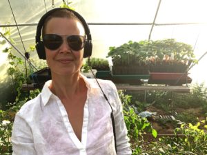 Sara står i vit skjorta i kvällsljus, solglasögon och hörlurar, i tunnelväxthuset.