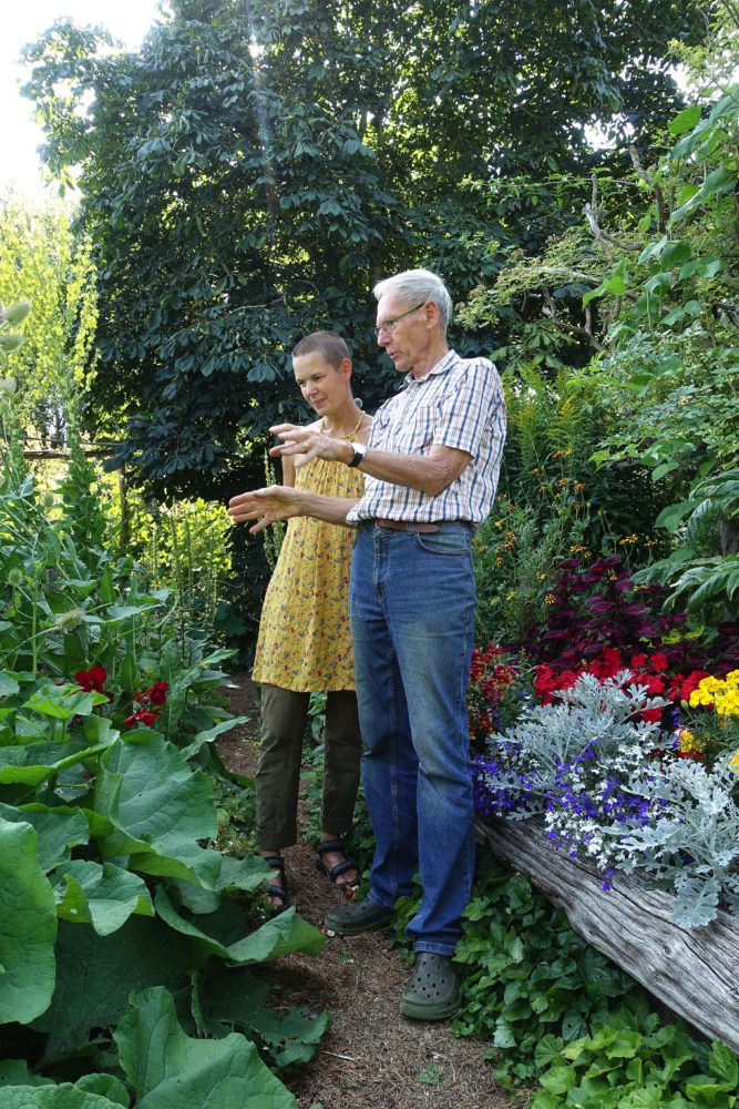 En kvinna i gul klänning och en äldre man i jeans och skjorta står i en trädgård och diskuterar.