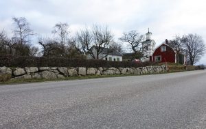 En litet rött hus längs vägen med vitt uthus och kyrka i bakgrunden