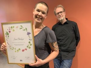 Två personer vid en röd vägg, en håller ett diplom.