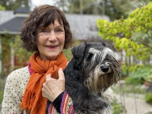 En kvinna med orange scarf står med en hund i famnen i sin trädgård.